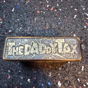 以下が含まれることがあります： 「THE DADDY TAX」の文字が施された、長方形の金属製のオブジェクト。文字は使い込まれたようなゴールドカラー。木目調の背景とゴールドカラーのフレームが特徴。背景はまだら模様の暗い表面。
