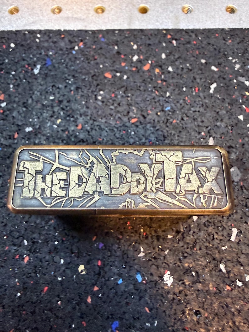 以下が含まれることがあります： 「THE DADDY TAX」の文字が施された、長方形の金属製のオブジェクト。文字は使い込まれたようなゴールドカラー。木目調の背景とゴールドカラーのフレームが特徴。背景はまだら模様の暗い表面。