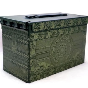 May include: An olive green metal box with detailed floral and geometric patterns. The box has a handle on the top and a latch closure. The design includes skulls and a central circular motif.