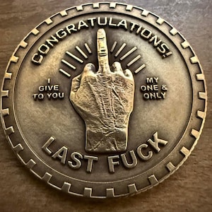 May include: A gold-colored metal coin with the words "Congratulations!" and "Last Fuck" around the edge. The center of the coin features a raised image of a middle finger with the words "I give to you" and "My one & only" on either side.