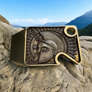 Puede incluir: Un abridor de botellas de metal dorado con forma de casco espartano con un diseño decorativo. El abridor de botellas está sobre una roca.