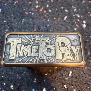 以下が含まれることがあります： 「TIME TO PAY」の文字がひび割れたような書体で刻まれた長方形の金属オブジェクト。金属はブロンズ仕上げで、文字は明るく対照的な色です。このオブジェクトはおそらく装飾品またはユニークな芸術作品です。