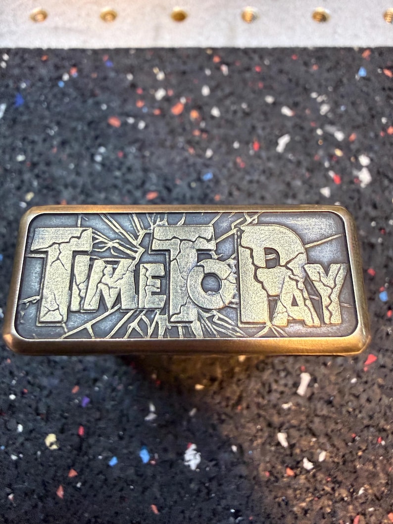 以下が含まれることがあります： 「TIME TO PAY」の文字がひび割れたような書体で刻まれた長方形の金属オブジェクト。金属はブロンズ仕上げで、文字は明るく対照的な色です。このオブジェクトはおそらく装飾品またはユニークな芸術作品です。