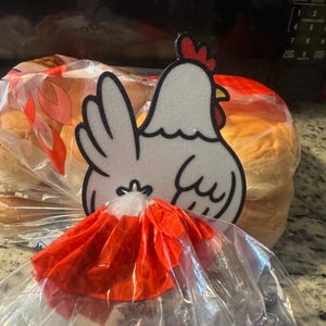 May include: A white and black chicken-shaped decoration with a red comb and a bright orange skirt, placed on top of a loaf of bread in a clear plastic bag. The bread is golden brown.