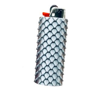 Puede incluir: Un encendedor plateado con una cubierta texturizada en forma de escamas. El encendedor tiene un bot&oacute;n de encendido rojo y una parte superior de metal. La cubierta est&aacute; hecha de peque&ntilde;as piezas superpuestas en forma de diamante, lo que le da una apariencia decorativa &uacute;nica.