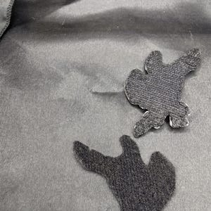 May include: Two black fabric patches with irregular, organic shapes are displayed against a smooth, dark gray textile background. The patches feature a textured, hook-and-loop fastener backing, showing a dense, fibrous surface structure.