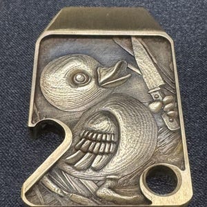 Può includere: Un apribottiglie rettangolare in metallo con un'incisione a rilievo dettagliata di un'anatra minacciata da un coltello. L'apribottiglie ha un apribottiglie e un piccolo foro circolare. Il metallo è color bronzo.
