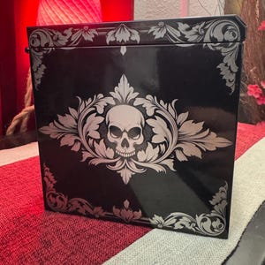 May include: Black box with a silver skull and floral design. The box has a glossy finish.
