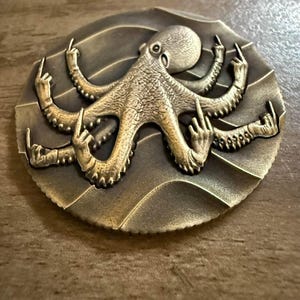 May include: A gold-coloured metal coin featuring a detailed sculpture of an octopus with all eight tentacles giving the middle finger. The octopus is set against a textured background of waves.