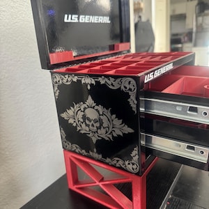Puede incluir: Una caja de joyer&iacute;a negra y roja con un dise&ntilde;o de calavera y el texto "U.S. GENERAL" en la parte delantera. La caja tiene dos cajones y un compartimento superior con varios compartimentos.