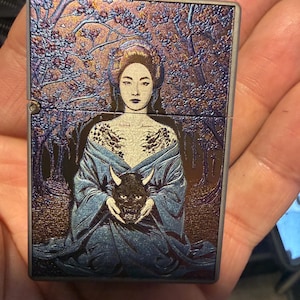 Puede incluir: Un encendedor metálico rectangular con acabado iridiscente y un grabado detallado de una persona con túnica azul que sostiene una máscara oscura con cuernos. El fondo muestra un bosque con árboles en tonos violeta y dorado.