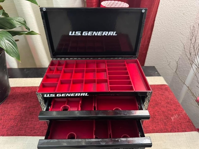 Puede incluir: Caja de joyer&iacute;a negra con compartimentos rojos y dos cajones extra&iacute;bles. La caja tiene las palabras "U.S. GENERAL" impresas en la parte delantera y superior.