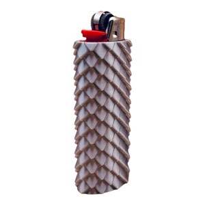 Puede incluir: Un encendedor decorativo con un patr&oacute;n texturizado en forma de escamas en tonos marrones y grises. El encendedor tiene un bot&oacute;n de encendido rojo y una parte superior de metal plateado.