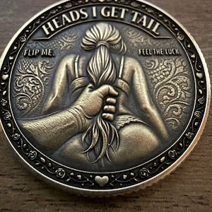 Peut inclure: Une pièce de monnaie ronde de style antique avec le texte "HEADS I GET TAIL" et "FLIP ME. FEEL THE LUCK." La pièce présente un relief d'une personne tirée par les cheveux. La pièce a une bordure décorative et un symbole de cœur.