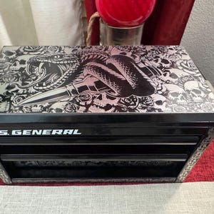 May include: Black metal toolbox with three drawers. The top drawer has a graphic of a snake and skulls with the text "U.S. GENERAL" on the front.