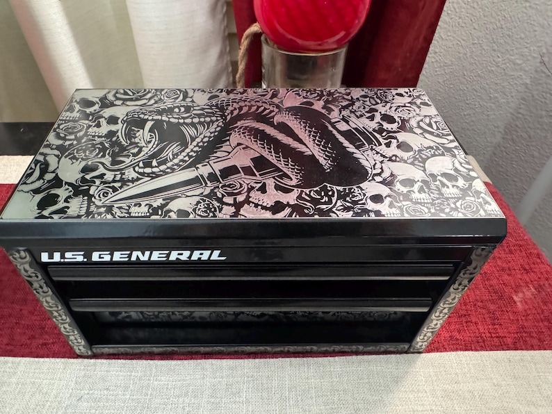 May include: Black metal toolbox with three drawers. The top drawer has a graphic of a snake and skulls with the text "U.S. GENERAL" on the front.