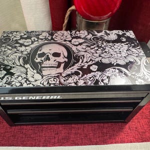 May include: Black metal toolbox with three drawers. The top drawer has a skull and floral design on the lid. The toolbox is labeled "U.S. GENERAL".