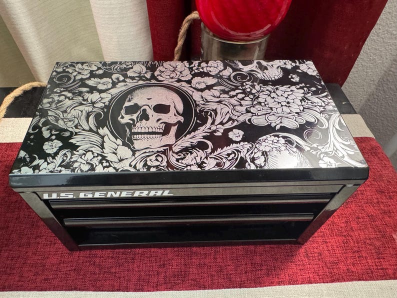 May include: Black metal toolbox with three drawers. The top drawer has a skull and floral design on the lid. The toolbox is labeled "U.S. GENERAL".