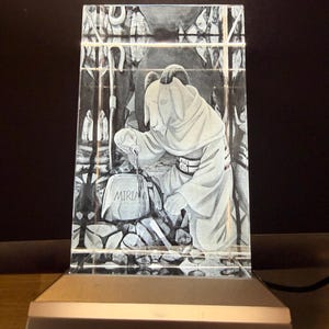 May include: A rectangular, clear glass block with an etched image of a robed figure kneeling at a tombstone. The tombstone reads "MIRIAM". The block is illuminated by a silver base with an on/off power button.