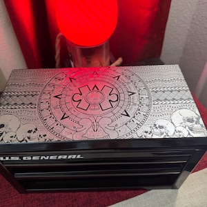 May include: Black metal toolbox with three drawers. The top drawer has a decorative design featuring a circular pattern with skulls and a stylized Aztec calendar. The toolbox is labeled "U.S. GENERAL".