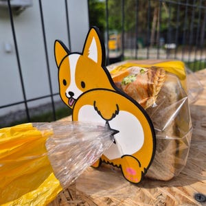 May include: A whimsical, orange and white corgi-shaped bread clip. The clip is attached to a yellow bread bag, with the corgi's rear end facing the viewer. The clip has a black outline and pink paw pads.