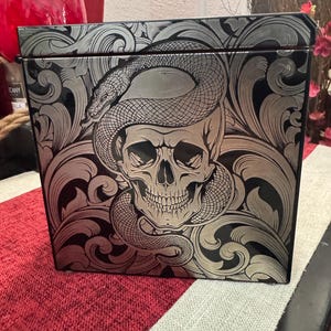 May include: A black box with a silver skull and snake design. The skull has a toothy grin and the snake is coiled around the skull's head. The design is intricate and detailed.