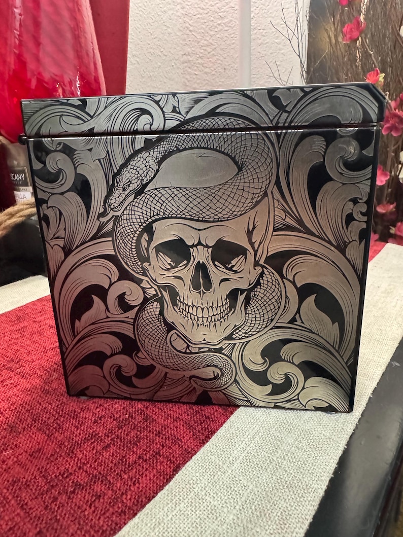 May include: A black box with a silver skull and snake design. The skull has a toothy grin and the snake is coiled around the skull's head. The design is intricate and detailed.