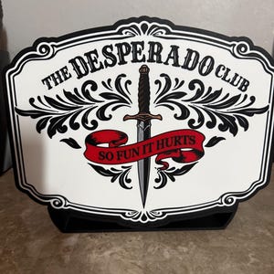 May include: A decorative sign with the text "THE DESPERADO CLUB" and "SO FUN IT HURTS". The sign features a black and white design with a dagger in the center and a red banner. The sign is displayed on a black stand.