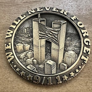 Puede incluir: Una moneda conmemorativa de color bronce que representa las Torres Gemelas con una bandera estadounidense. La moneda está inscrita con "WE WILL NEVER FORGET" y "9/11". La moneda tiene un fondo detallado de paisaje urbano.