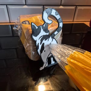 May include: A black and white cat-shaped bread clip, holding a clear plastic bread bag closed. The cat has a spotted pattern and a curved tail. The bread is in a yellow and clear plastic bag.