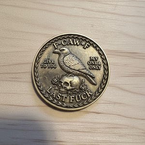 May include: A round, antique-style metal coin with a detailed design. The coin features a crow perched atop a skull with roses, and the text "F-CAW-F", "I GIVE TO YOU", "MY ONE & ONLY", and "LAST FUCK".