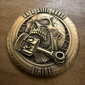 Puede incluir: Moneda de color bronce con un diseño detallado de calavera y pistón. La moneda tiene el texto "ON A STEEL HORSE I RIDE" alrededor del borde. La moneda tiene un aspecto texturizado y envejecido.