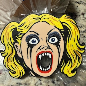 May include: A cartoon-style image of a woman's face with yellow hair, blue eye shadow, and black tears. The mouth is open, revealing sharp white teeth. The image has a black outline.