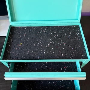 May include: An open, turquoise metal toolbox with two pull-out drawers. The interior of the box and drawers are lined with a speckled black material. The box is on a dark surface.
