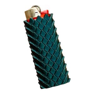 Puede incluir: Un encendedor verde azulado con una textura tejida. El encendedor tiene una parte superior de metal dorado y un bot&oacute;n de encendido rojo. La textura tejida es un patr&oacute;n de diamantes repetido.