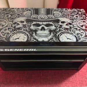 以下が含まれることがあります： 3つの引き出しが付いた黒いツールチェスト。上部にはシルクハットをかぶったスカルが描かれ、歯車と時計に囲まれています。「U.S. GENERAL」の文字が前面にプリントされています。