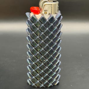 Puede incluir: Una funda de encendedor negra e iridiscente con un patr&oacute;n geom&eacute;trico de diamantes. El encendedor tiene un bot&oacute;n rojo y una parte superior de metal plateado. La funda est&aacute; dise&ntilde;ada para un encendedor est&aacute;ndar y a&ntilde;ade un toque decorativo.