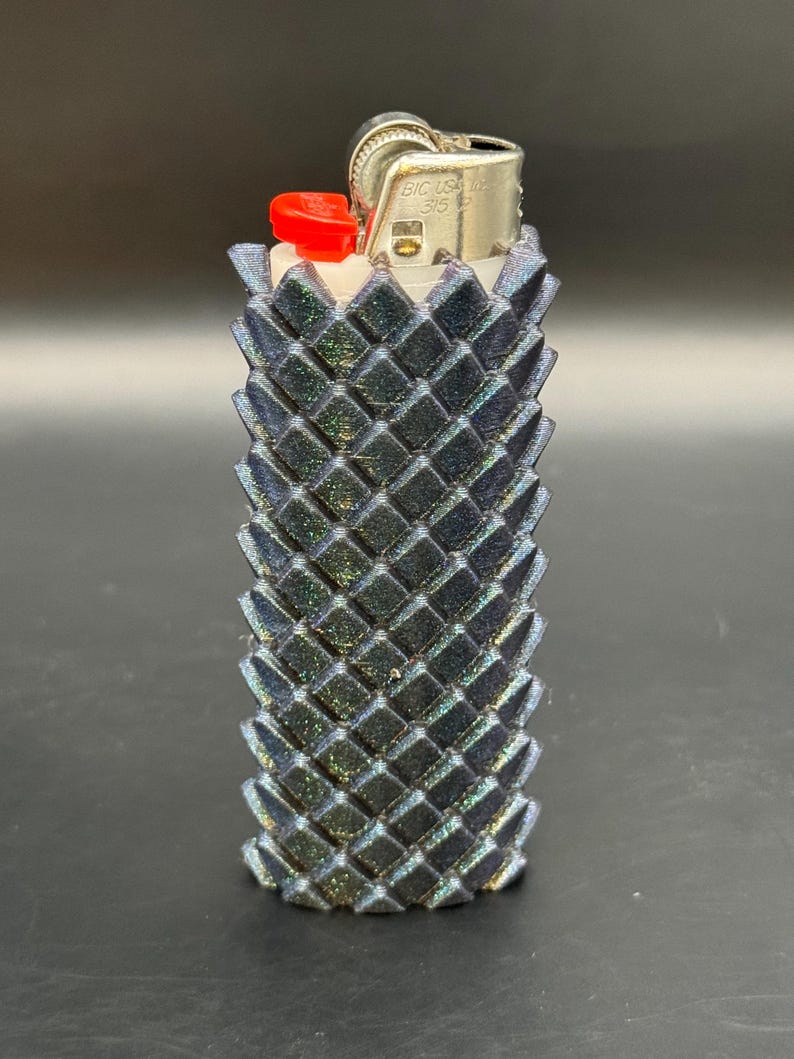 Puede incluir: Una funda de encendedor negra e iridiscente con un patr&oacute;n geom&eacute;trico de diamantes. El encendedor tiene un bot&oacute;n rojo y una parte superior de metal plateado. La funda est&aacute; dise&ntilde;ada para un encendedor est&aacute;ndar y a&ntilde;ade un toque decorativo.