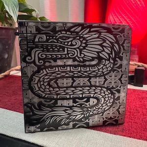 May include: A black box with a silver and black design of a dragon with intricate details. The dragon is curled around a geometric pattern.