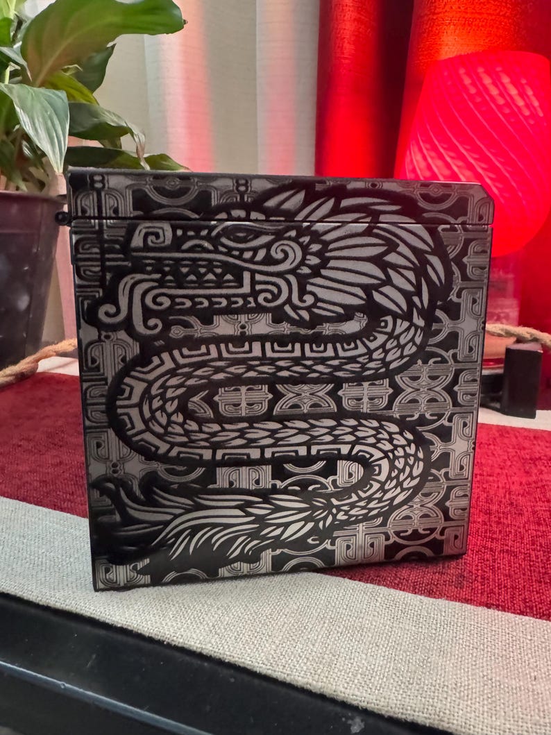 May include: A black box with a silver and black design of a dragon with intricate details. The dragon is curled around a geometric pattern.