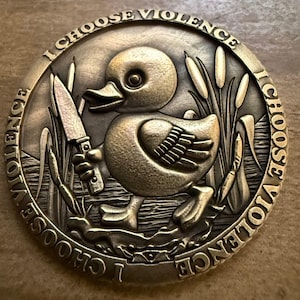 May include: A bronze coin with a duck holding a knife, set amongst reeds. The coin is inscribed with the words "I CHOOSE VIOLENCE". The duck has a determined expression.