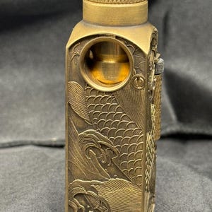 Pode incluir: Um isqueiro retangular de cor latão antigo com intrincadas gravuras de ondas e escamas de peixe. O isqueiro apresenta um topo texturizado e uma abertura circular, mostrando um design vintage detalhado.