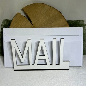 May include: White wooden sign with the word "MAIL" in large, cutout letters. The sign is sitting on top of a white envelope.