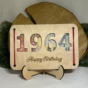 May include: A wooden card with the year 1964 cut out and filled with money. The card has a red string attached to the top and bottom and says "Happy Birthday" on the bottom. The card is sitting on a wooden stand.