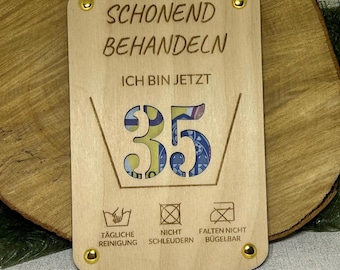May include: Wooden plaque with the text "SCHONEND BEHANDELN ICH BIN JETZT 35" and care symbols. The number 35 is filled with a colorful pattern. The plaque is held together with four gold-colored rivets.
