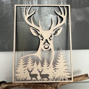 May include: Wooden wall art depicting a deer head with prominent antlers, crafted from light-coloured wood. The design incorporates a forest scene with two deer and trees. The artwork is framed in a rectangular shape.