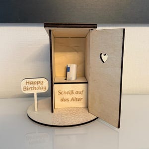 Könnte beinhalten: Ein helles Holzobjekt in Form eines Plumpsklos als Geldgeschenk mit einer aufklappbaren Tür und einem herzförmigen Ausschnitt. Die Vorderseite ist mit 'Scheiß auf das Alter' graviert und ein Schild trägt die Aufschrift 'Happy Birthday'.