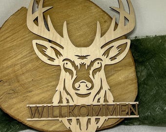 May include: Wooden deer head wall art with the word "WILLKOMMEN" cut out below. The deer head is light brown and the sign is mounted on a round, light brown wooden base. The antlers are detailed and the deer's face is intricately carved.