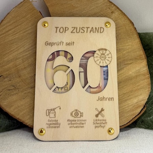 Puede incluir: Una placa rectangular de madera clara con el texto grabado TOP ZUSTAND y 60 Jahren. El número 60 está recortado, revelando billetes detrás. Incluye una pegatina de inspección circular y tres iconos con texto en alemán.