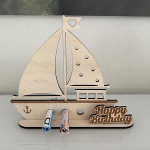 Könnte beinhalten: Geschenkehalter in Form eines Holzsegelbootes mit herzförmiger Flagge. Der Halter hat ein Ankerdesign und hält aufgerollte Geldscheine. Die Worte "Happy Birthday" sind auf der Basis angezeigt.